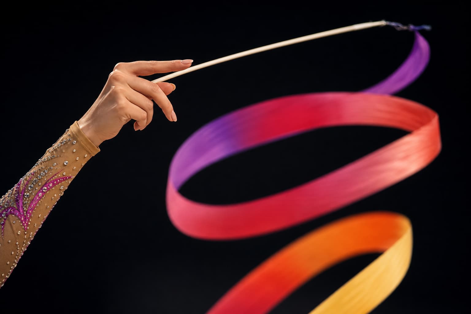 Une main de gymnaste rythmique tient un bâton de ruban, le ruban coloré formant une spirale dans les airs avec un flou de mouvement sur fond sombre.