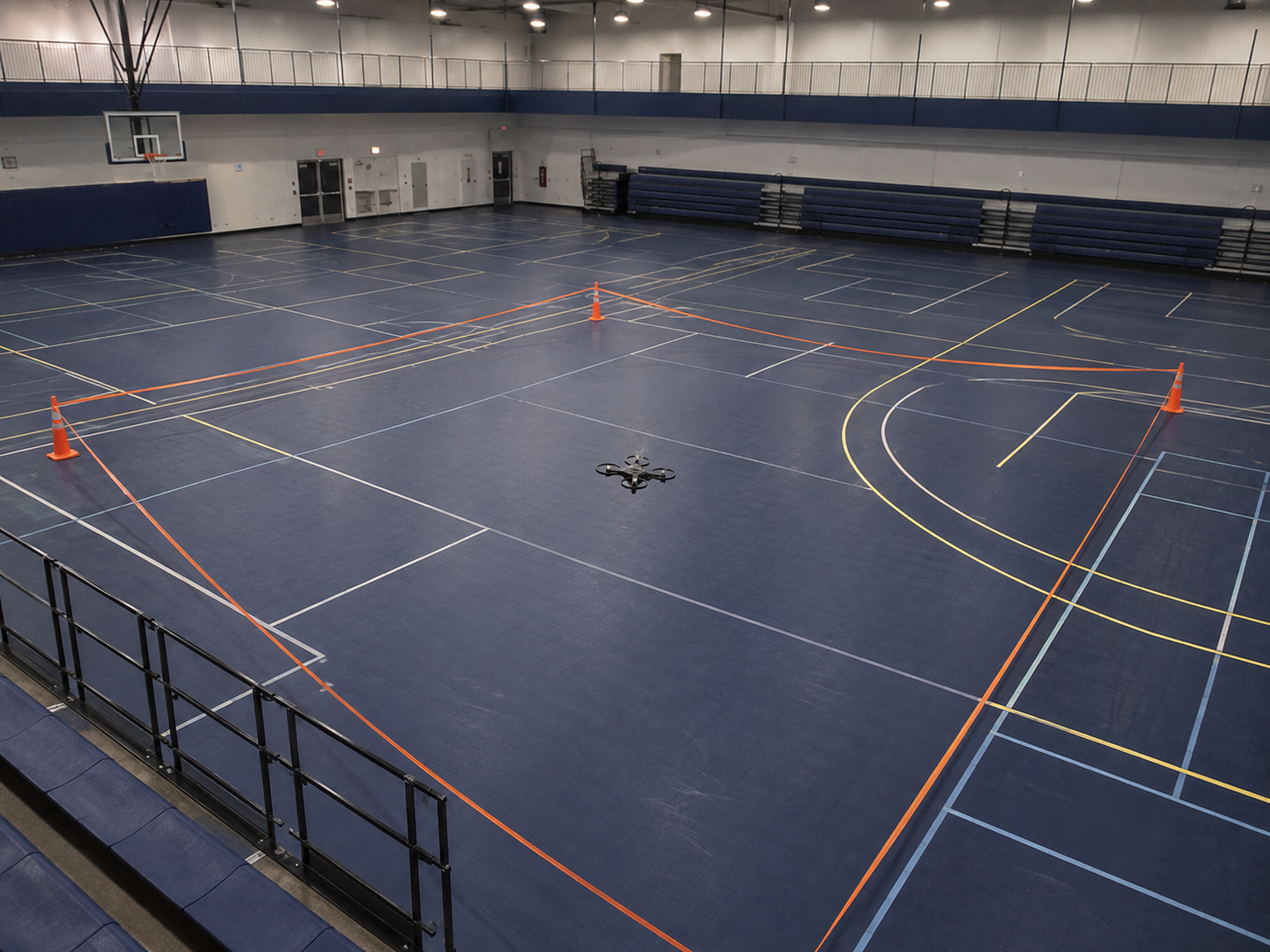 Petit drone générique en vol bas dans une zone délimitée par des cônes et un ruban orange, gymnase vide, aucun visage.