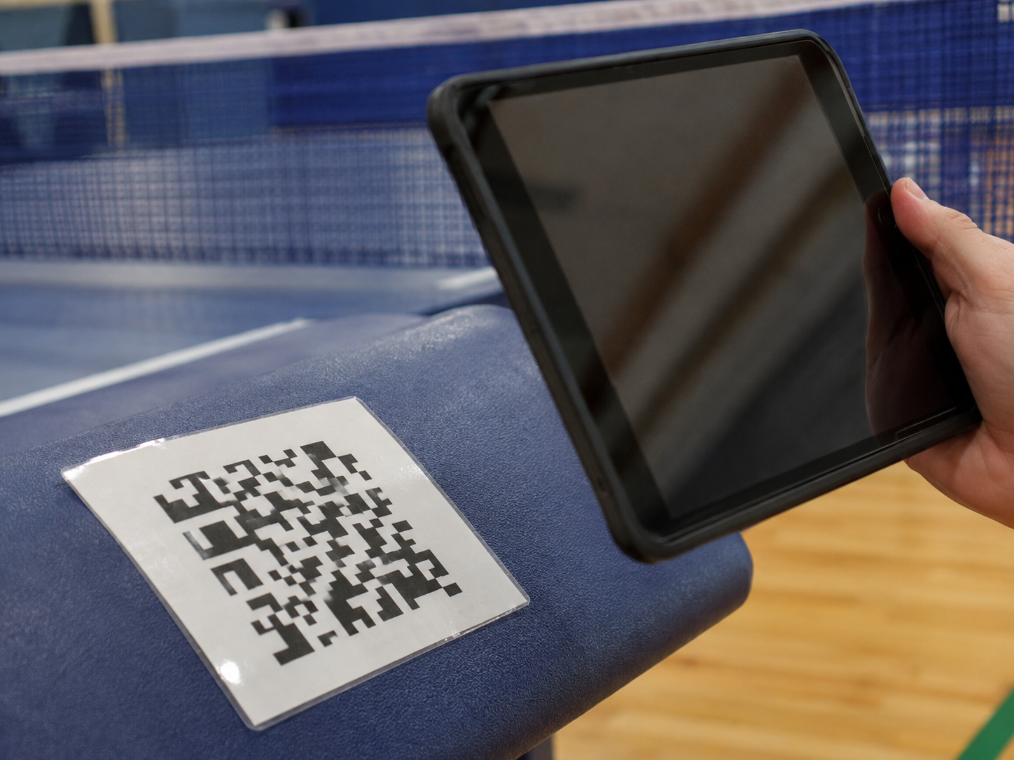 Illustration : fiche d’atelier plastifiée avec un code QR décoratif non opérationnel, tablette tenue pour le scan, filet de tennis de table flou.